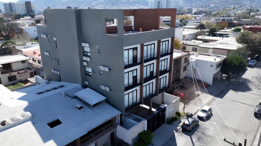 Aerial view of a modern four-story gray apartment building with black-framed windows on a residential street, with balconies and hills in the background.