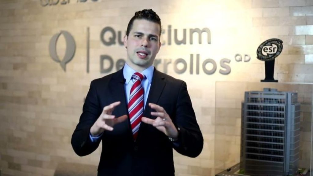 Man in a dark suit and red striped tie gesturing while speaking in a modern office, with a company sign on the wall behind him and a trophy on a pedestal.