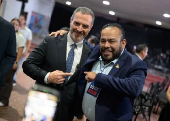 Two suited men smiling and posing together at an event, one with his arm around the other’s shoulder while they point at each other in agreement.