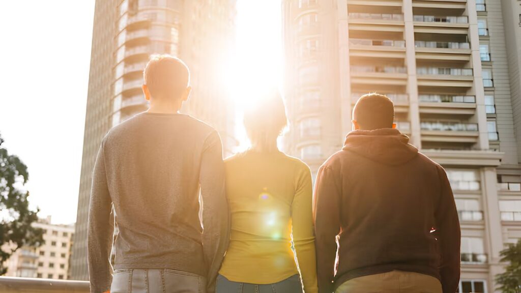 vida-barrio-urbana - LA VISION 360 Tres amigos están de espaldas a la cámara en un paisaje urbano bañado por el sol, con la luz filtrándose entre los altos edificios que se alzan tras ellos.