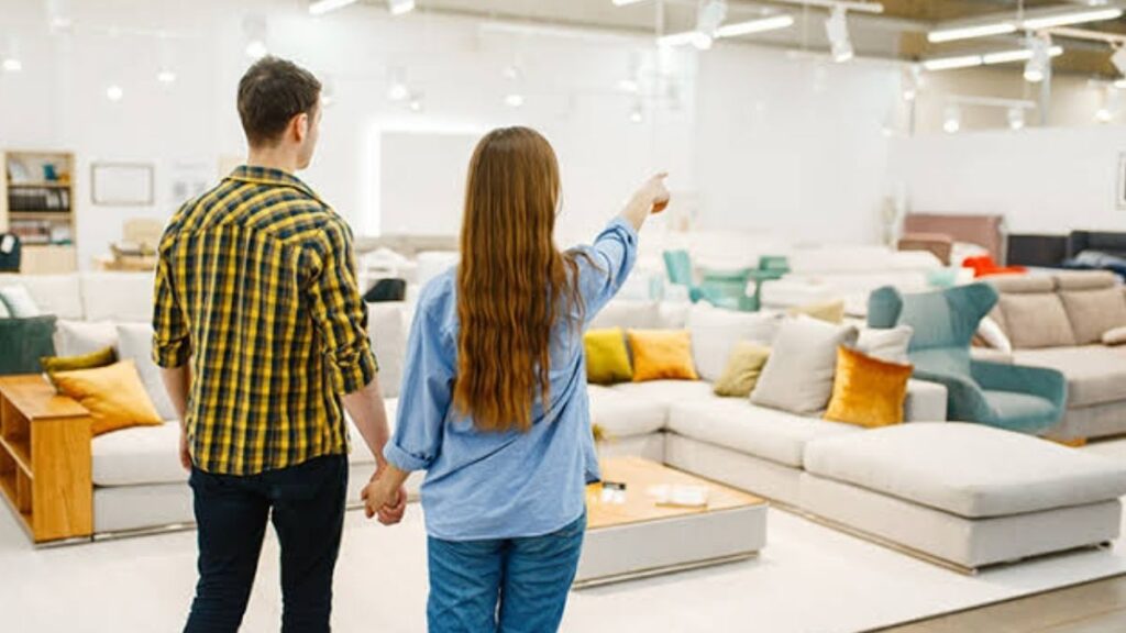 vision-360-gastos-ocultos-comprar-casajpg - LA VISION 360 Couple holding hands walks through a bright furniture showroom, the woman pointing at a piece of furniture ahead of them.